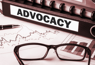 A stack of binders and notebooks is on a table. One says "ADVOCACY" on the spine. Also on the table is a paper with line graphs and bar charts, an ink pen, and a pair of dark-rimmed glasses.