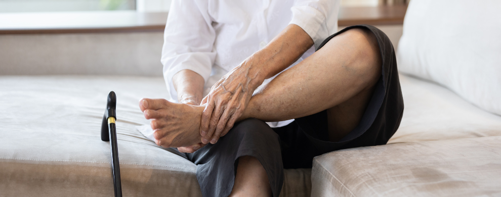 A man massages his ankle as he sits on a bed with one leg hanging over the side and the other crossed over it with the ankle resting on the knee.