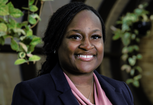 Alondra Dorsey is standing in a hallway with hanging plants. She has long black braids, is smiling, and wearing a pink blouse under a navy blue jacket.