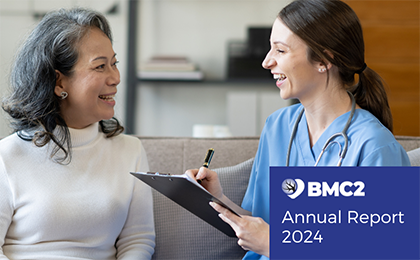 A young white health care provider smalls while speaking with a patient and writing notes on a clip board. She is smiling, wears blue scrubs and a stethoscope around her neck and has long dark hair pulled back in a ponytail. Her patient, an older Asian woman with shoulder-length salt and pepper hair, wearing a white turtleneck sweater, smiles back.
